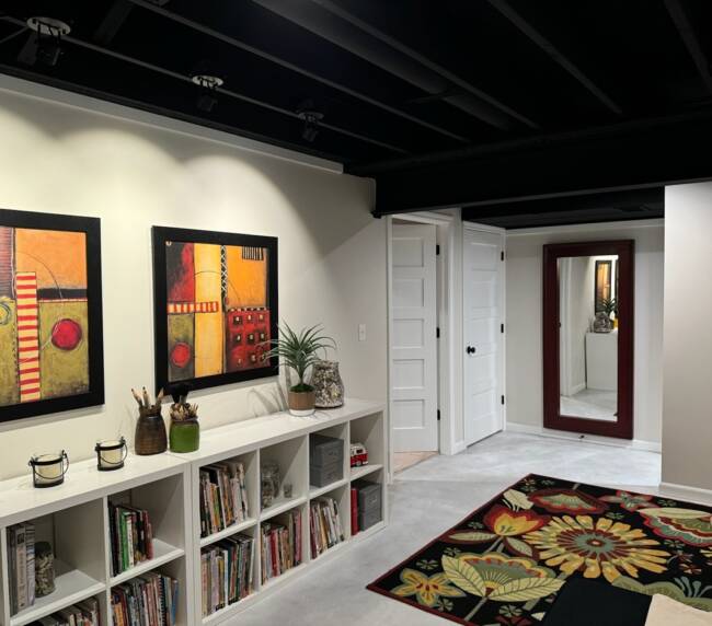Room with a black ceiling, abstract art on the wall, white bookshelves filled with books, a colorful floral rug, and a large red-framed mirror