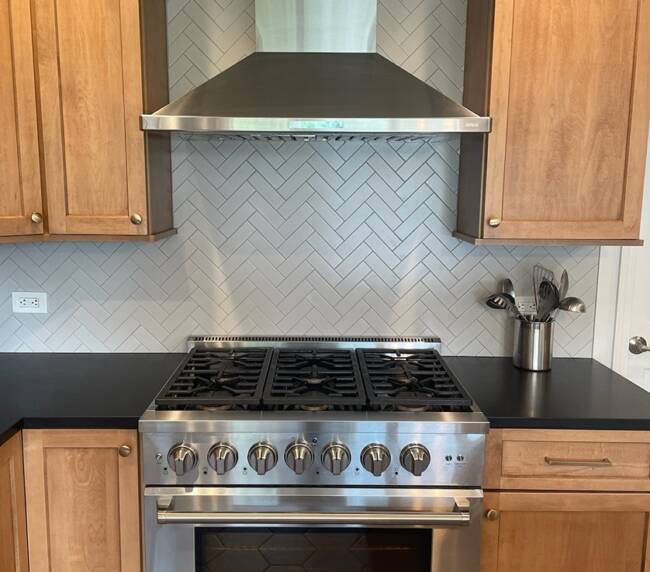 kitchen with a stainless steel range and hood, black countertop, light wood cabinets, and a herringbone tile backsplash
