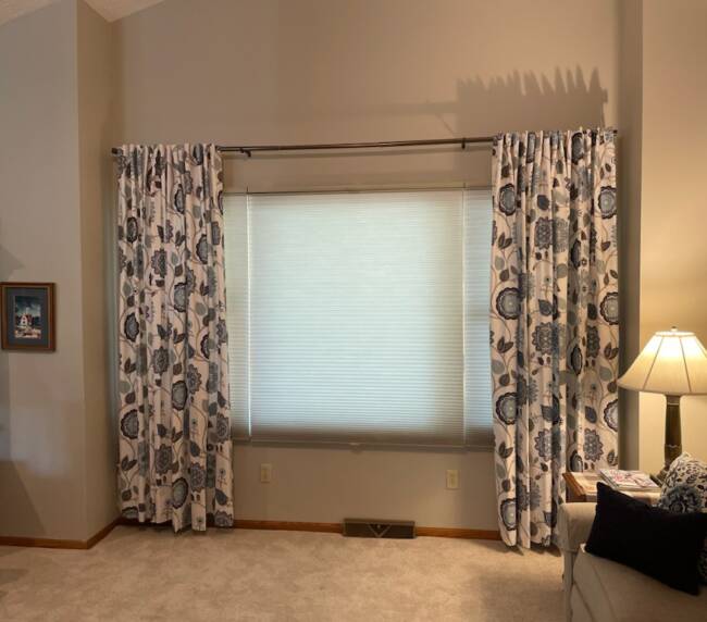 Room with tall patterned curtains flanking a window with cellular shades, and beige carpeted flooring