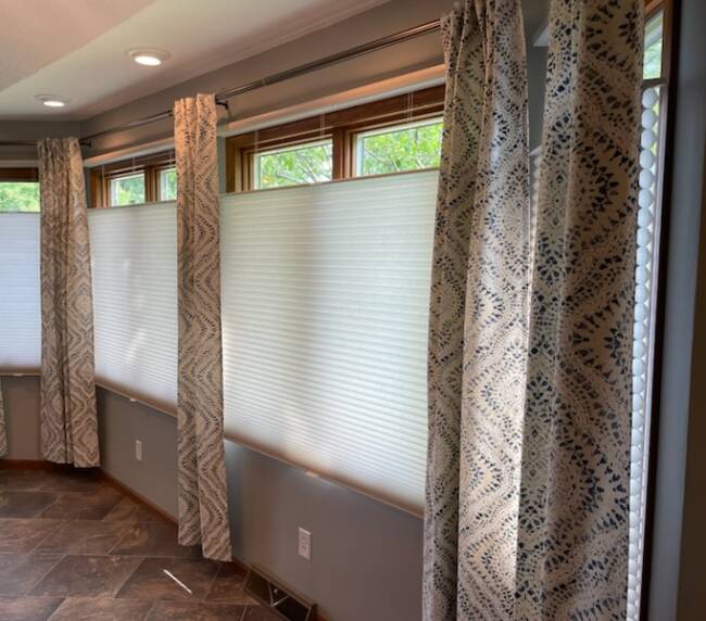 Room with large windows covered by patterned curtains and cellular shades, and dark tiled flooring