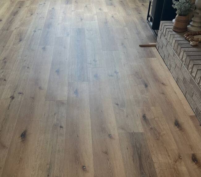 wood plank flooring in a living area, leading towards a window with blinds, and showing a portion of a brick fireplace