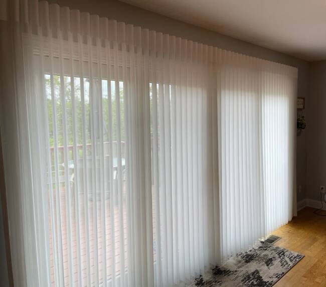 Room with large sliding glass doors covered by white vertical blinds, a view of a deck outside, and light wood flooring