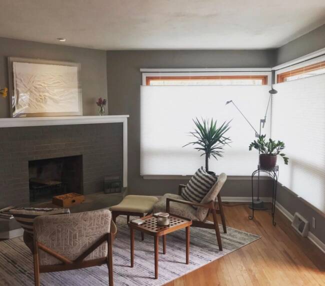 Cozy living room with a gray tiled fireplace, modern furniture, wooden flooring, and large windows with white shades