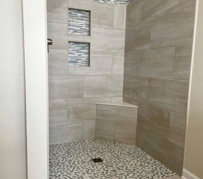 Shower with large beige tiles and an accent border of small mosaic tiles. The floor features pebble-like mosaic tiles